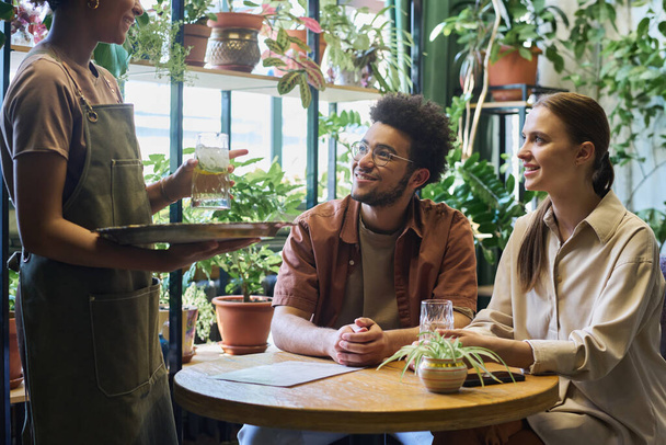 Gelukkige jonge man en vrouw zitten aan tafel tegen groene planten in cafe en kijken naar serveerster met dienblad en glas verfrissende limonade - Foto, afbeelding