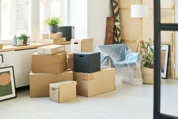 Stacks of packed cardboard boxes standing on the floor by windowsill and in the corner of living room with rolled carpets and domestic plant - Photo, Image