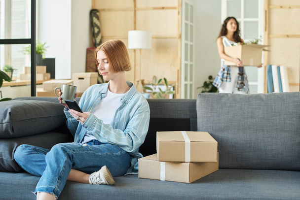 Jonge vrouw in denim casualwear zittend op zachte comfortabele bank in woonkamer van nieuw appartement, met thee en sms 'en in smartphone - Foto, afbeelding