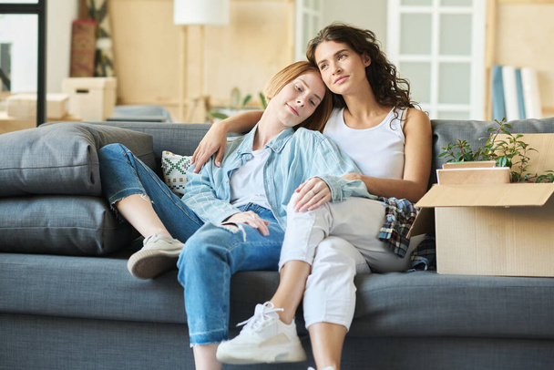 Zwei glückliche junge Freundinnen in Freizeitkleidung sitzen auf einer bequemen Couch im Wohnzimmer und genießen die Ruhe nach dem Umzug in eine neue Wohnung - Foto, Bild