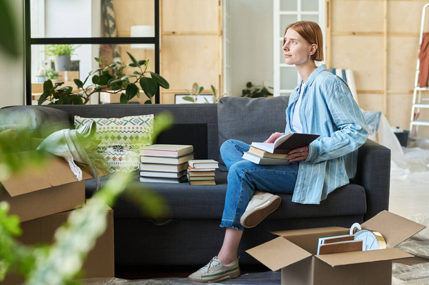 Jeune femme avec pile de livres sur ses genoux assis sur un canapé confortable et doux dans le salon et se reposer après avoir déménagé dans un nouvel appartement - Photo, image