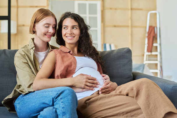 Two happy young women sitting on couch in front of camera after relocation while one of them embracing her pregnant girlfriend - Photo, Image