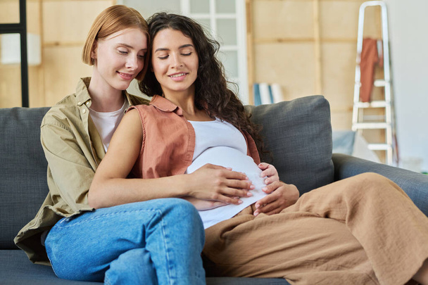 Happy young brunette pregnant woman and her affectionate girlfriend sitting on couch and enjoying rest after relocation to new apartment - Photo, Image