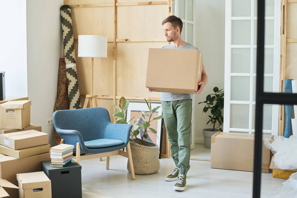 Young male loader carrying big cardboard package while moving along spacious living room with furniture and packed things during relocation - Photo, Image