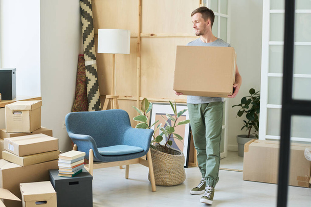 Junger Mann in Freizeitkleidung trägt große schwere Kiste und betrachtet Stapel anderer Pakete, die am Wohnzimmerfenster auf dem Boden stehen - Foto, Bild