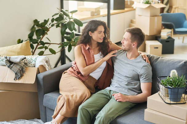 Junge Frau blickt ihren Mann an, während sie ihn umarmt und auf weicher bequemer Couch im geräumigen Wohnzimmer etwas diskutiert - Foto, Bild