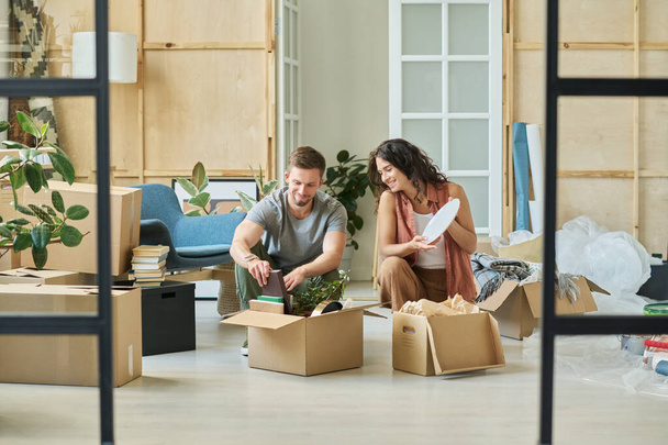 Feliz joven mujer y su marido desempacando cajas de cartón con utensilios de cocina, libros, plantas domésticas verdes, reloj y otras cosas - Foto, imagen