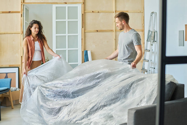 Giovane coppia divano di copertura con imballaggio cellophane protettivo durante la preparazione di mobili per la ristrutturazione e la pittura delle pareti del soggiorno - Foto, immagini