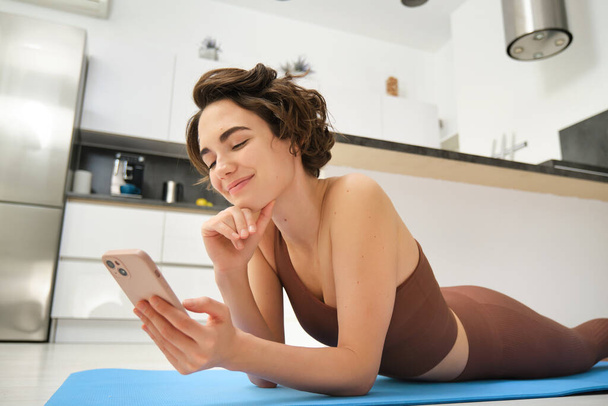 Sprawność i dobre samopoczucie online. Portret młodej kobiety leżącej na macie do jogi, patrzącej na aplikację na smartfona, patrzącej na wskazówki treningowe, trenującej w domu w stroju aktywnym. - Zdjęcie, obraz