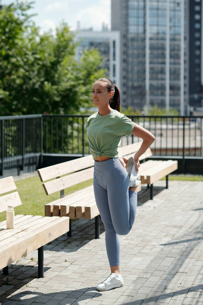Μακρύ πλάνο της νεαρής δραστήριας μελαχρινής γυναίκας σε αθλητικά ρούχα άσκηση εξωτερική, ενώ στέκεται δίπλα στον πάγκο κατά πράσινα δέντρα και κτίρια - Φωτογραφία, εικόνα