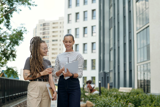 Gelukkige jonge zakenvrouw met drank tonen document met financiële gegevens aan collega terwijl beide lopen langs hoge moderne gebouwen - Foto, afbeelding