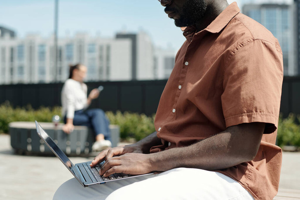 Ritagliato colpo di giovane analista afroamericano maschio premendo i tasti della tastiera del computer portatile mentre seduto di fronte alla fotocamera in ambiente urbano - Foto, immagini
