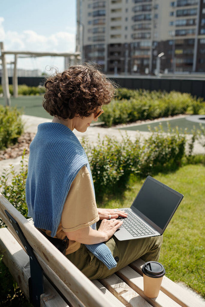 Seitenansicht einer jungen brünetten Geschäftsfrau mit Laptop auf Knien, die auf einer Bank sitzt und sich Online-Videos ansieht oder Nachrichten durchsieht - Foto, Bild