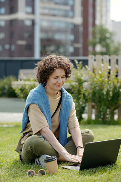 Счастливый молодой сотрудник или фрилансер с ноутбуком, сидящий на зеленом коврике в летний день и работающий над новым проектом или анализирующий онлайн-данные - Фото, изображение