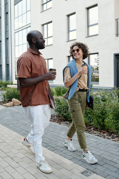 Feliz joven pareja intercultural o colegas con tazas de café caminando por edificios modernos en la calle y discutiendo algo - Foto, imagen