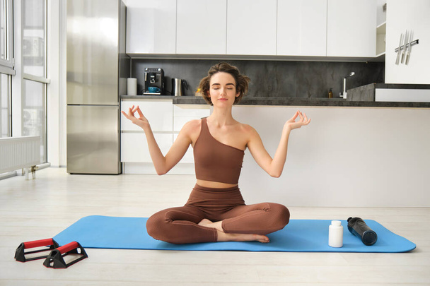 Ritratto di giovane donna fitness, atleta donna pratica yoga da casa, seduta in posa di loto con viso rilassato, utilizzando tappetino di gomma in cucina per l'allenamento. Sport e concetto di benessere - Foto, immagini