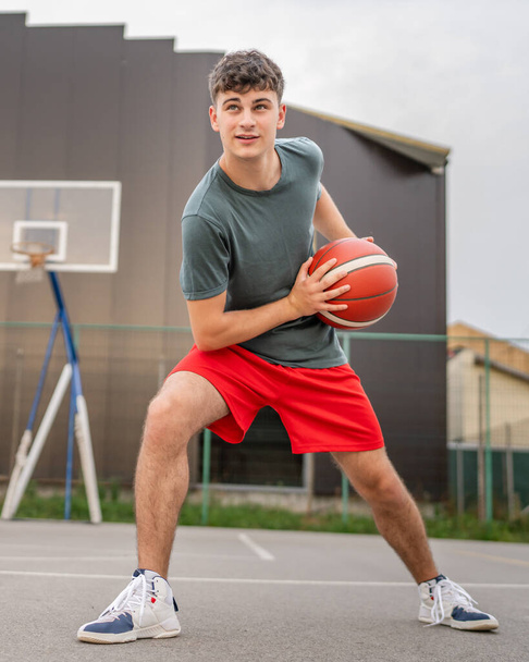 Un adolescent caucasien mâle caucasien jeune homme debout sur le terrain de basket-ball avec balle dans la soirée prêt à jouer copie jeu espace personne réelle - Photo, image