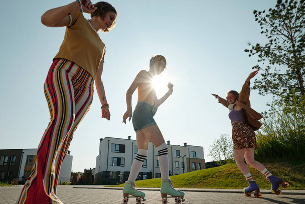 Три безтурботні дівчата в повсякденному одязі і роликових ковзанах насолоджуються проведенням часу на літніх вихідних, в той час як одна з них стоїть на тлі сяючого сонця - Фото, зображення