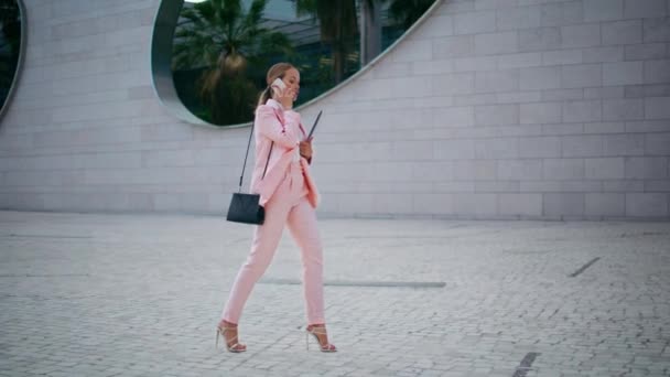 Hermosa señora jefa llamando caminando por la calle adoquines en elegante traje rosa. Empresaria exitosa teniendo conversación de trabajo por teléfono móvil va en la oficina. Mujer confiada hablando smartphone. - Metraje, vídeo
