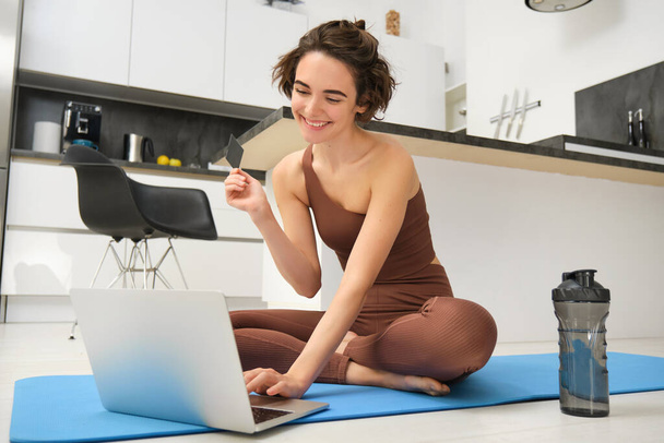 Image of young fitness girl, female athlete at home, workout on kitchen floor, holding credit card with laptop, purchasing online training session, gym application for home workout. - Photo, Image
