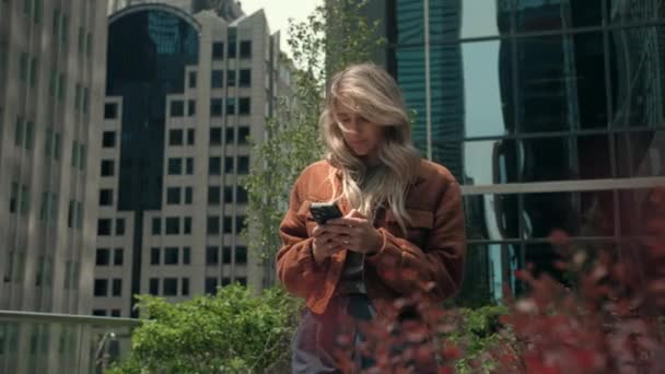 Mujer rubia joven utilizando el teléfono de navegación por Internet en el fondo de la ciudad.La señora usa chaqueta naranja navegar por la web con mensajes de escritura de teléfonos inteligentes al aire libre.Gente adicta al teléfono celular. Imágenes de alta calidad 4k - Imágenes, Vídeo