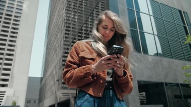 Молода блондинка використовує телефон, що переглядає Інтернет на фоні міста. Леді носить помаранчеву куртку, що серфінгує Інтернет зі смартфоном, який друкує повідомлення на відкритому повітрі. Високоякісні 4k кадри - Кадри, відео