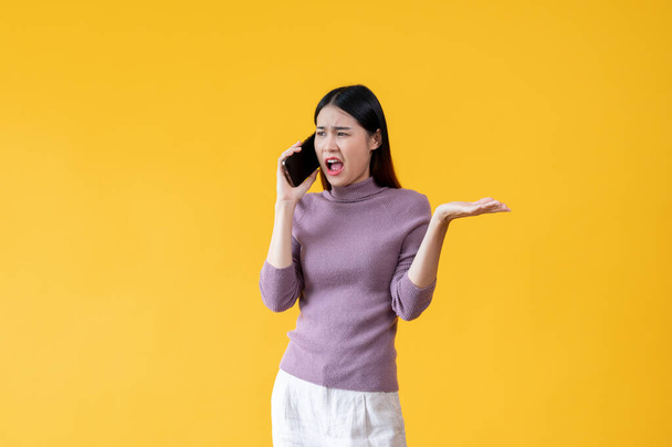 Een woedende en ongelukkige Aziatische vrouw praat agressief aan de telefoon, klaagt over iemand aan de telefoon, voelt zich ontevreden. geïsoleerde gele achtergrond - Foto, afbeelding