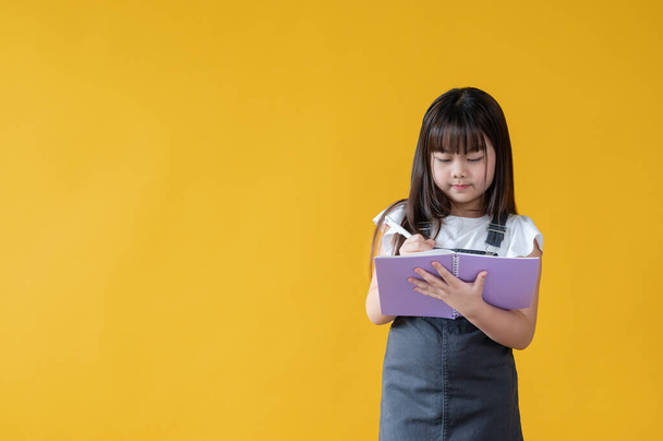 Симпатичная маленькая азиатка фокусируется на том, чтобы написать что-то в своей книге или сделать домашнее задание, стоя на изолированном жёлтом фоне. Школьница, студентка, дети, школьник - Фото, изображение