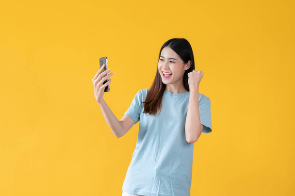 Una joven asiática feliz y emocionada está mirando la pantalla de su teléfono inteligente con una cara feliz y mostrando su puño hacia arriba, de pie sobre un fondo amarillo aislado. - Foto, Imagen