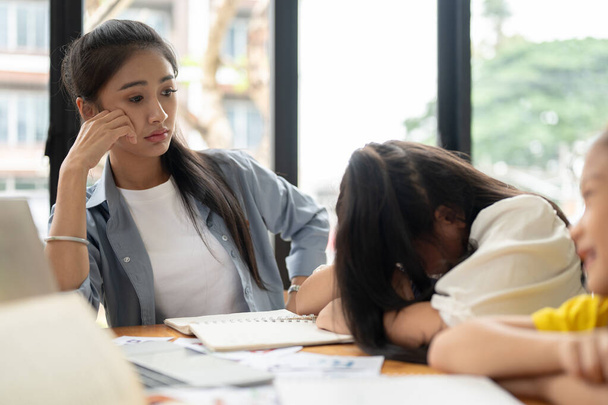 Een geïrriteerde en overstuur Aziatische lerares en een koppige, slaperige studente zitten aan een tafel in de klas. onderwijsprobleem voor kinderen - Foto, afbeelding