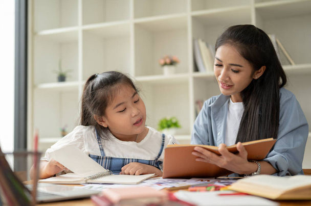 Een vriendelijke Aziatische moeder helpt met huiswerk en geeft Engels aan haar dochtertje thuis in het weekend. mam en dochter, student en leraar, babysitten - Foto, afbeelding