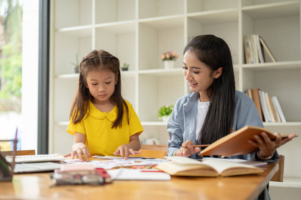 Una adorable niña preescolar asiática se está centrando en estudiar tarjetas de vocabulario inglés con un amable profesor privado en casa. preescolar, jardín de infantes, estudiante de primaria - Foto, imagen