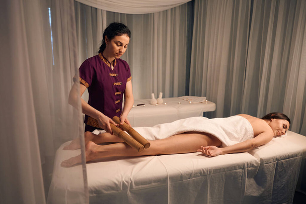 Женщина-клиент лежит склонной, пока массажистка катит пару бамбуковых тростей над мышцами ее голени - Фото, изображение