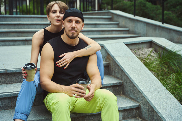 Beautiful couple in casual clothes with coffee cup looking at camera and sitting on stairs in park - Fotografie, Obrázek
