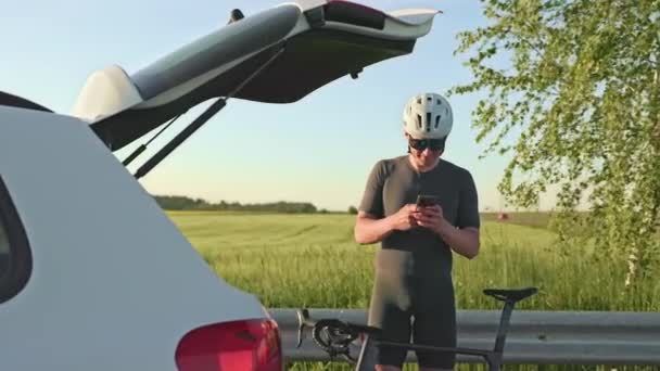 Young fit man with sports gearing utilizing smartphone while waiting with racing cycle near open car boot on sunny day. Delighted athlete checking email after finishing outdoor bike riding workout. - Footage, Video