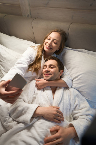 Beautiful woman hugging her handsome husband by neck making selfie on smartphone, couple resting and having fun lying in bed in warm soft bathrobes - Photo, Image