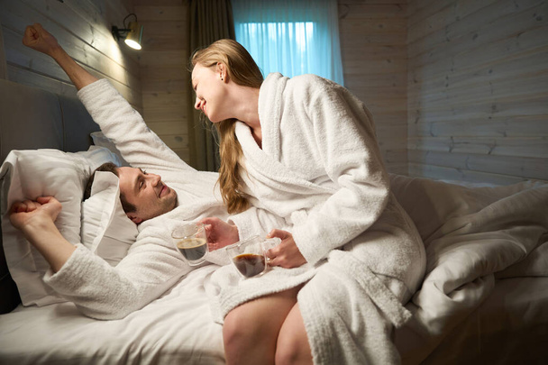 vrouw wakker haar man met aroma van hete verse koffie ze brengt, paar in de liefde genieten van ochtend routine, tijd doorbrengen in romantische sfeer - Foto, afbeelding