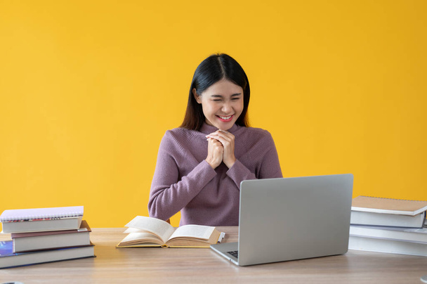 Een mooie Aziatische vrouw kijkt naar haar laptop scherm met een opgewonden en gelukkig gezicht, zittend aan haar bureau tegen een geïsoleerde gele achtergrond. - Foto, afbeelding