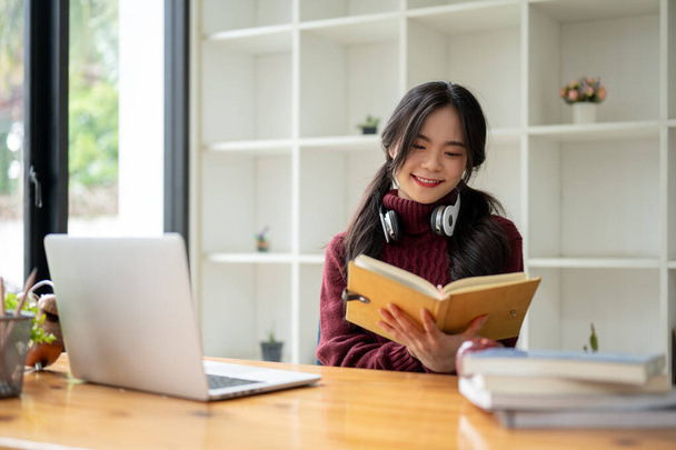 Een vrolijke en charmante jonge Aziatische studente in een knusse rode trui leest een boek aan een tafel in de coffeeshop. - Foto, afbeelding