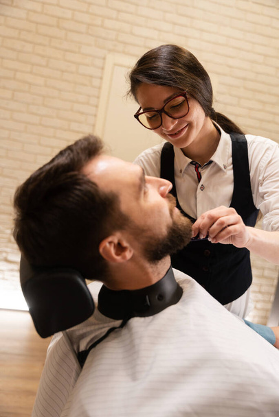 Linda hembra peluquero corta una barba a un cliente, el hombre se encuentra en una silla cómoda - Foto, Imagen
