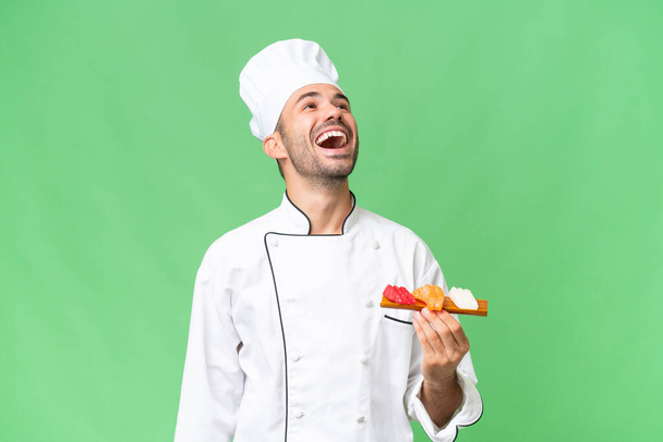 Giovane chef caucasico che tiene un sushi su sfondo isolato ridendo - Foto, immagini