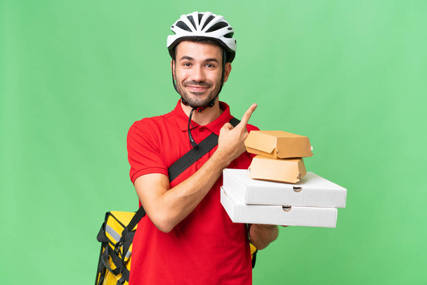 Young handsome caucasian man with thermal backpack and holding takeaway food over isolated background pointing to the side to present a product - Photo, Image