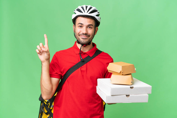 Young handsome caucasian man with thermal backpack and holding takeaway food over isolated background showing and lifting a finger in sign of the best - Photo, Image