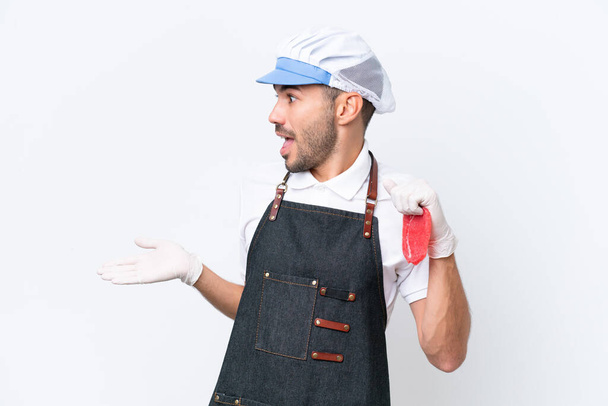 Boucher homme caucasien portant un tablier et servir de la viande fraîche coupée sur fond blanc isolé avec expression surprise tout en regardant côté - Photo, image
