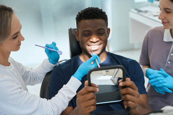 Afrikai amerikai ügyfél ül a fogászati szék és a gazdaság tükör, nézi fogorvos mutatja, hogyan kezeli, és tiszta, fogászati protézisek - Fotó, kép