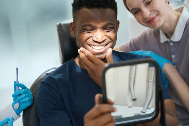 Νοσοκόμα ενθαρρύνοντας τον αρσενικό πελάτη που κοιτάζει τα δόντια του στον καθρέφτη και χαμογελά, ικανοποιημένος με την υγεία και την ποιότητα, οδοντιατρική προσθετική - Φωτογραφία, εικόνα
