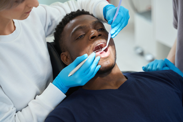 Koncentrált női fogorvos ellenőrzése egészség afro-amerikai ügyfél fogak segítségével száj tükör és sarló szonda, megérintve minden fog, hogy ellenőrizze zománc minősége - Fotó, kép