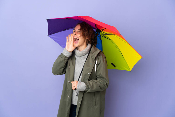 Anglická žena drží deštník izolovaný na purpurovém pozadí křičí s ústy dokořán otevřenými do strany - Fotografie, Obrázek