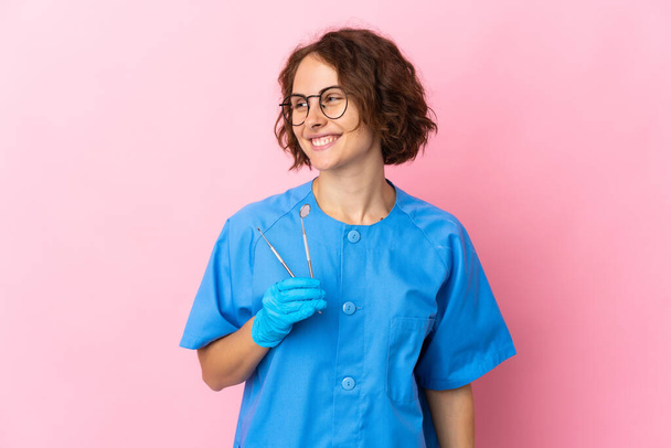 Mujer Inglés dentista sosteniendo herramientas sobre aislado en rosa fondo mirando a un lado y sonriendo - Foto, Imagen