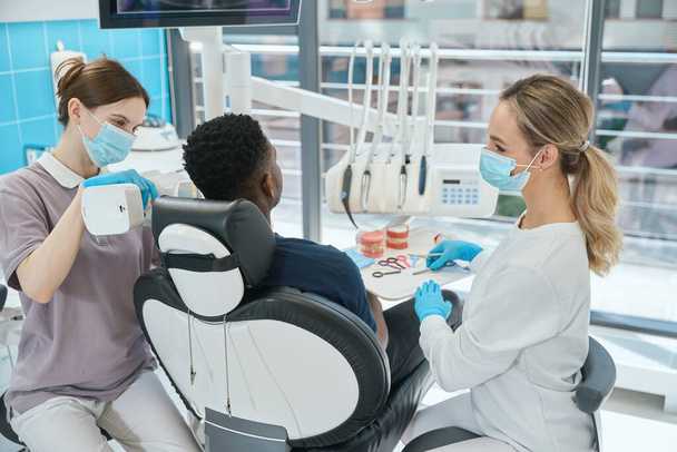 Kadın stomatolog bağlanmış ve bekliyor. Afro-Amerikalı hasta dişlerinin röntgenini çekiyor. Portatif röntgen kamerası, estetik dişçilik... - Fotoğraf, Görsel